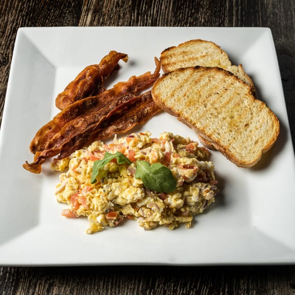 Scrambled eggs with tomatoes and onions, crispy bacon, and grilled toast on a white plate.