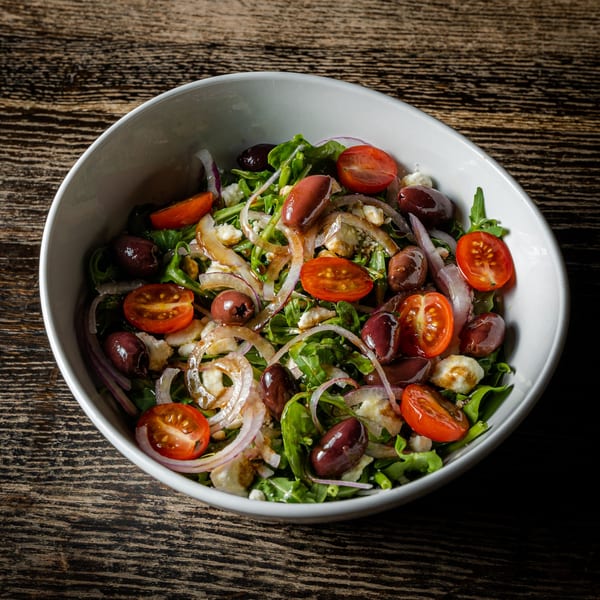 A vibrant salad with arugula, cherry tomatoes, olives, red onions, and feta cheese in a white bowl.