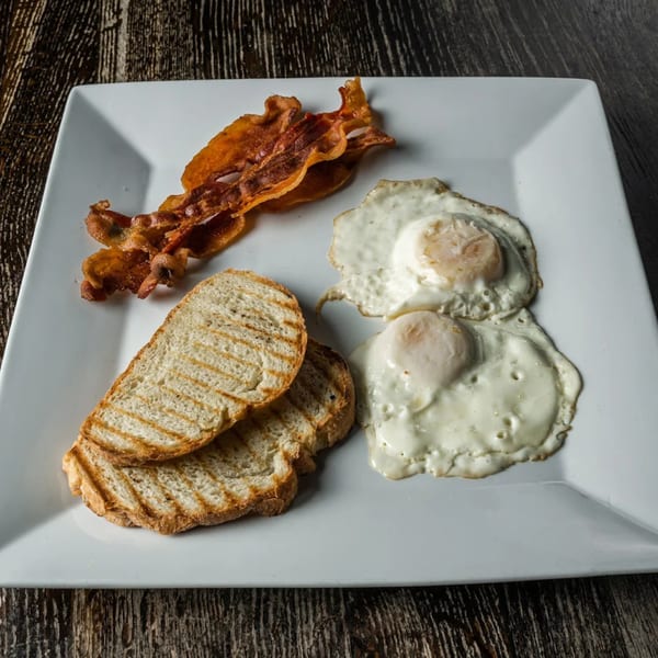A breakfast plate with crispy bacon, two fried eggs, and grilled toast on a white square plate.