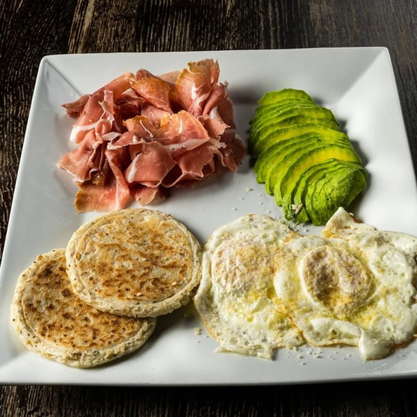 A Caffe Gourmet breakfast plate with prosciutto, sliced avocado, two arepas, and fried eggs.