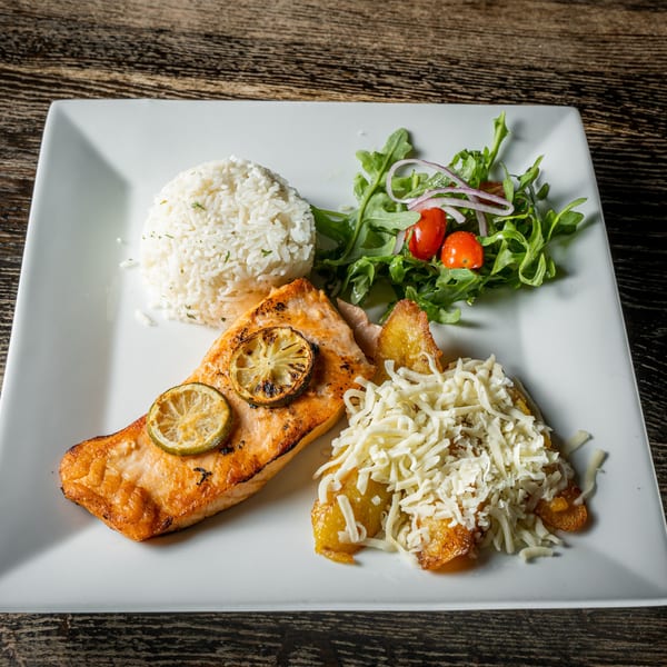 Grilled salmon with lime, rice, salad, and fried plantains topped with shredded cheese on a white plate.